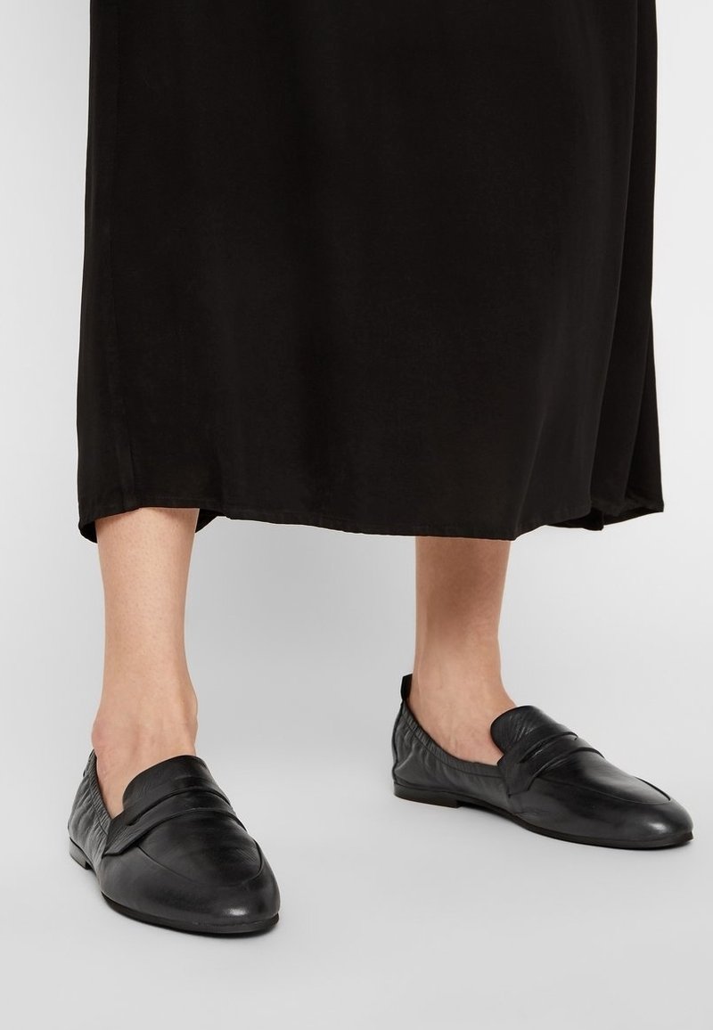 Zapatos negros de piel de tipo slip-on con un acabado suave, que presentan pliegues decorativos y una suela plana. El diseño es angular con acentos texturizados.