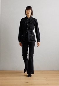 Svart denimjakke med belte i midjen og to brystlommer, kombinert med flagrende svarte jeans. Jevn tekstur, enkel design og metallknapper.