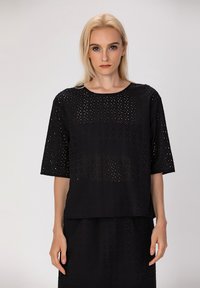 Haut noir à manches courtes avec motif en œillets ; tissu texturé ; encolure rond ; coupe ample ; associé à une jupe noire assortie.
