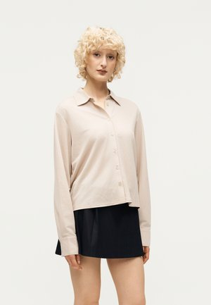 Νέος με σγουρά ξανθά μαλλιά που φοράει μπεζ πουκάμισο με κουμπιά και μαύρη φούστα, στέκεται μπροστά σε ένα απλό φωτεινό φόντο.