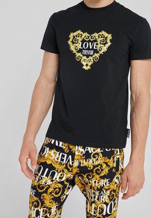 Czarny t-shirt z żółtym ozdobnym sercem i napisem "LOVE VERSACE JEANS COUTURE" zestawiony z czarnymi spodniami z żółtymi wzorami barokowymi i białym napisem.