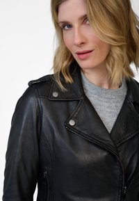 Schwarze Leder-Motorradjacke mit silbernen Druckknöpfen, Reißverschlusstaschen und einem gekerbten Kragen, über einem hellgrauen Pullover.