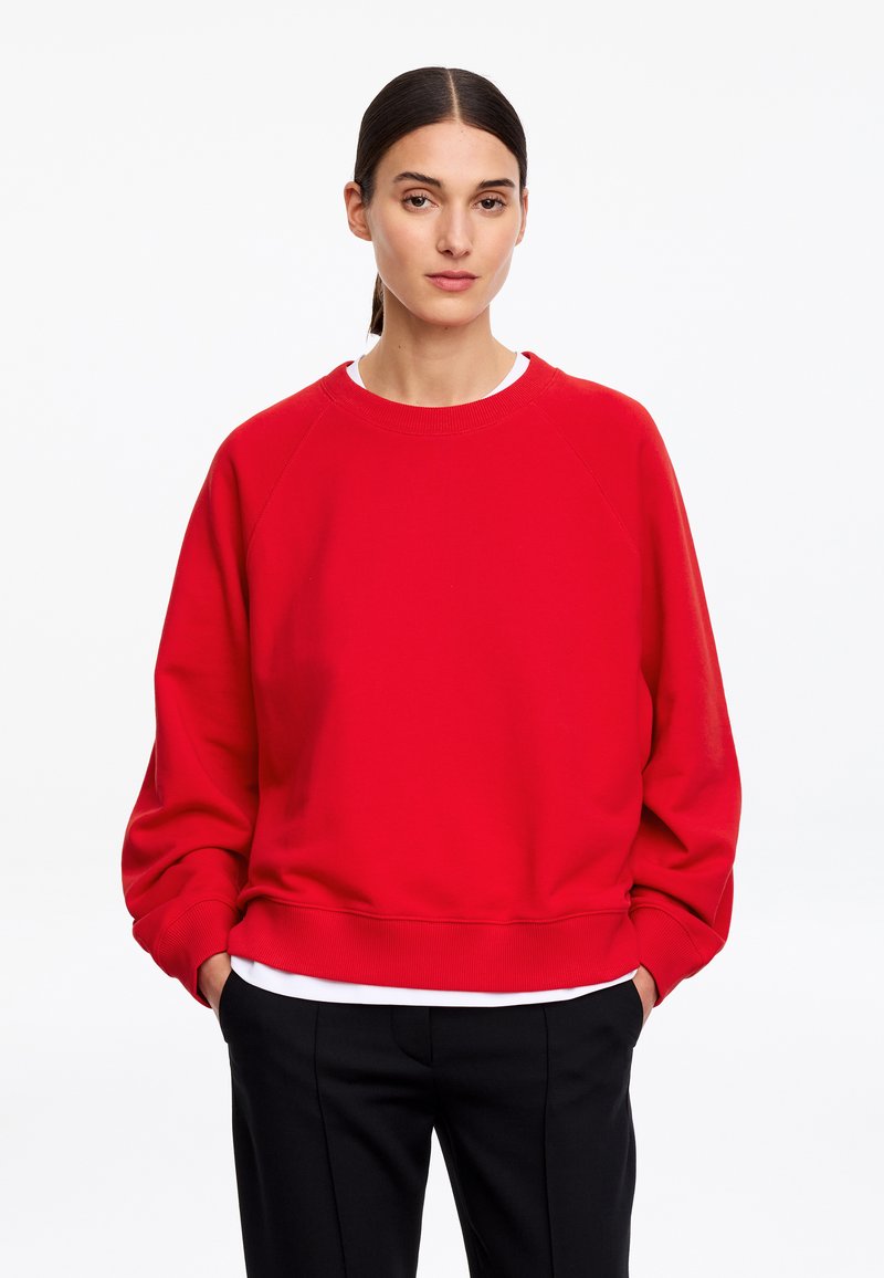 Femme aux cheveux foncés en queue de cheval, portant un sweat rouge ample, un t-shirt blanc et un pantalon noir, debout, les mains dans les poches.