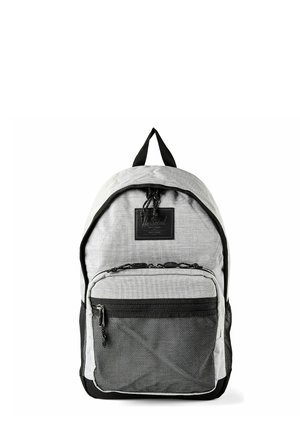 Sac à dos en tissu gris clair avec fermeture éclair noire, poche avant en maille et poignée supérieure, posé contre un fond blanc.