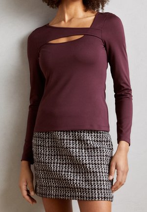 Femme portant un haut bordeaux à manches longues avec un détail découpé et une mini-jupe texturée noir et blanc.