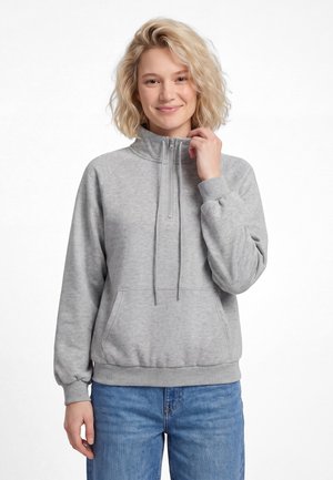 Ung kvinde med krøllet blondt hår smilende, iført en lysegrå quarter-zip sweatshirt og blå jeans, stående foran en hvid baggrund.