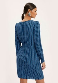 Niebieska sukienka w wzory z długimi rękawami, okrągłym dekoltem i dopasowanym krojem, zdobiona abstrakcyjnym geometrycznym nadrukiem w ciemniejszym odcieniu niebieskiego.