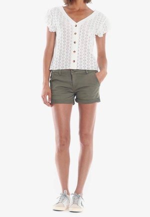 Kvinde iført en hvid knapfast kortærmet top, olivengrønne shorts med opbukket kant og hvide sneakers, stående med den ene hånd i lommen.