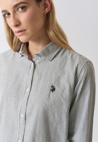Camicia a righe con bottoni in grigio e bianco, caratterizzata da un colletto appuntito e un piccolo logo ricamato sul petto sinistro. Tessuto morbido al tatto.