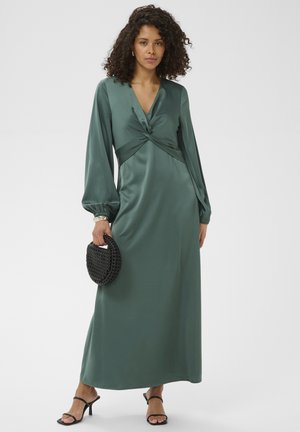 Femme aux cheveux bouclés portant une robe longue à manches longues en satin vert, tenant un sac à main tissé noir, et portant des sandales à talons noires.