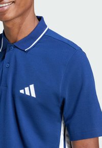 Blauwe polo met een witte kraag en gestreepte accenten, met een wit logo op de borst. Gemaakt van geweven katoen.