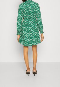 Groene bloemenjurk met lange mouwen, een kraag en een aangesloten taille. Heeft een uitlopende zoom en delicate bloempatronen. G gedragen met zwarte hakken.