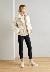 Chaqueta beige acolchada con cuello alto, combinada con vaqueros ajustados negros y botas cortas crema con herrajes decorativos y suela texturizada.