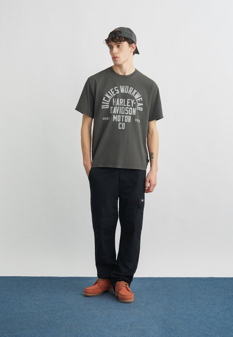 Giovane uomo con t-shirt grafica Harley Davidson grigio scuro, pantaloni cargo neri, stivali color tan e berretto grigio indossato al contrario, in piedi su pavimento blu.