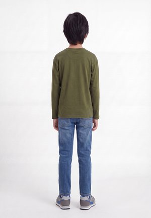 Niño de espaldas, vestido con una camiseta de manga larga verde oliva, vaqueros azules y zapatillas grises sobre un fondo blanco.