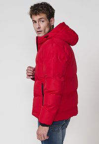 Chaqueta acolchada roja con capucha, Front con cremallera y puños elásticos. Presenta una textura suave con un patrón de acolchado horizontal.