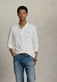 Νεαρός άνδρας στέκεται με το ένα χέρι στην τσέπη, φορώντας λευκό πουκάμισο με μακριά μανίκια και μπλε τζιν, μπροστά σε απλό γκρι φόντο.