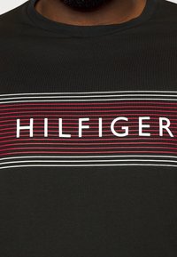 Musta t-paita, jossa on vaakasuorat punaiset ja valkoiset raidat, ja keskellä on paksuilla valkoisilla kirjaimilla teksti "HILFIGER".