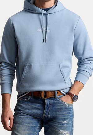 Mand iført lyseblå hættetrøje med forlomme, brun læderrem, blå jeans og armbåndsur, hånd i lommen, torso og arme synlige.