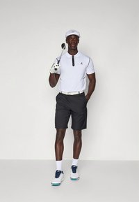 Fehér golfpóló fekete cipzárral, fekete rövidnadrággal párosítva. A ruházathoz fehér sapka, fehér zokni és kék díszítésű golfcipő tartozik.