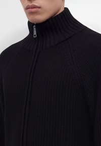 Gros plan d'une personne portant un pull en tricot côtelé noir zippé avec un col montant fermé jusqu'en haut.