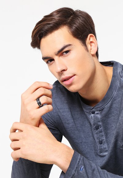 Maglietta henley a maniche lunghe blu navy con una patta a tre bottoni. Anello nero sulla mano, tonalità di pelle chiara, sfondo neutro.