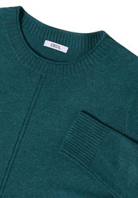 Maglione lavorato a maglia in teal con scollatura rotonda, polsini a coste e taschino sul petto. La trama del tessuto sembra morbida e leggermente melangiata. Etichetta del marchio visibile.