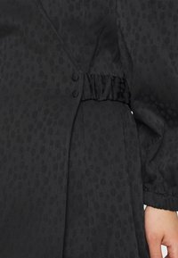 Svart, texturerad klänning med ett subtilt prickmönster, en elastisk midja och två dekorativa knappar på sidan. Långa, samlade ärmar.