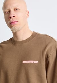 Sudadera de algodón marrón con cuello redondo. Presenta un pequeño logo rosa en el lado izquierdo del pecho. Textura suave con mangas en set-in.