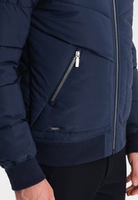 Námořnická pufrová bunda s prošívaným designem, zipovými bočními kapsami a ribovanými manžetami. Vyrobena z syntetického materiálu s hladkou texturou.