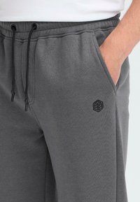 Grå joggesbukser laget av myk stoff har en elastisk midjebånd med justerbare snorer og en subtil svart logo på sidelommen.