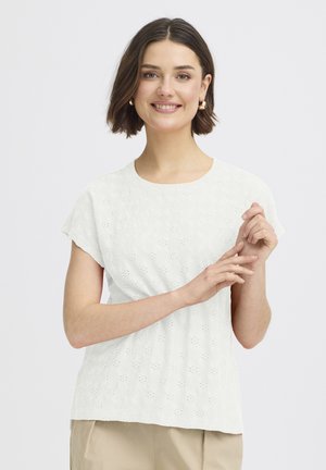 Noorpruunide lühikeste juustega noor naine, kellel on kullast rõngakujulised kõrvarõngad, valge silmaaugulise mustriga top ja beežid püksid, naeratades ja kerged käed kokku seotud.