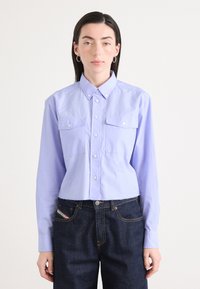 Camicia corta button-up di un leggero colore viola con due tasche sul petto e colletto a punta, abbinata a jeans denim scuri. Texture del tessuto liscia.