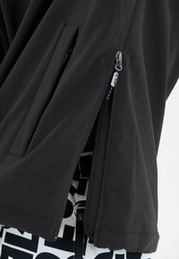 Sort lynlås sportsjakke med struktureret stof, sidelommer og afslappet pasform, med et skjult designelement under.