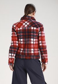 Rød og marineblå plaidjakke med et tekstureret, fuzzy materiale, der har hvide detaljer og en krave, set fra bagsiden.