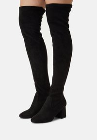 Botas de ante negras por encima de la rodilla con un tacón grueso y textura suave. Presenta puntera redondeada y detalle en los paneles laterales para un diseño adicional.