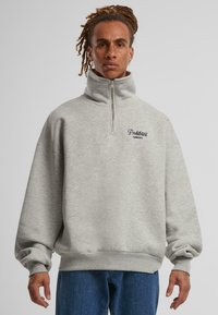 Sudadera gris claro con cuello alto, cierre de media cremallera y puños acanalados. Presenta un logo bordado de "Prendas Prohibidas" en el pecho.