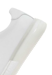 Baskets blanches avec une tige en synthétique lisse, un col rond et une semelle en caoutchouc translucide. Détail de panneau latéral texturé visible.