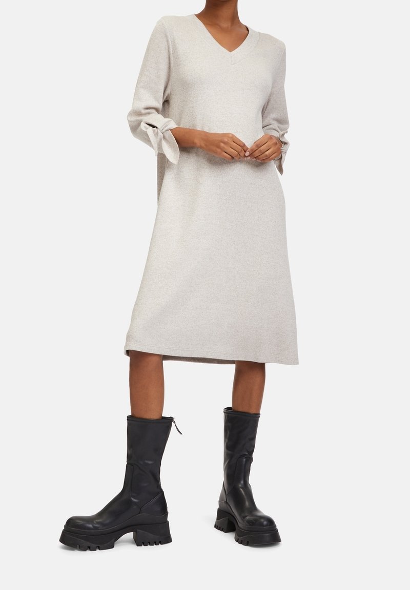 Femme portant une robe midi beige à col en V avec des manches 3/4 nouées et des bottes noires à semelle épaisse, se tenant devant un fond blanc.
