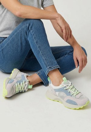 Osoba sedící v tureckém sedu, oblečená v modrých džínách a pastelových teniskách se světlezelenými podrážkami a tkaničkami na světle šedém pozadí.