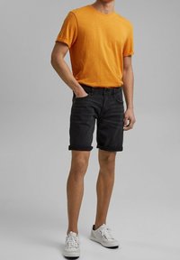 Pomarańczowa koszulka z podwiniętymi rękawami, zestawiona z czarnymi dżinsowymi szortami. Szorty mają podwinięty brzeg. Obuwie: białe sneakersy.