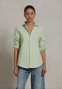 Chemise à boutons verte clair avec col et manches retroussées, fabriquée en tissu doux. Présente un petit logo sur le côté gauche de la poitrine.