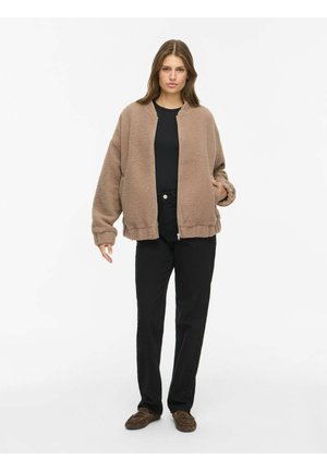 Femme debout portant une veste polaire beige zippée, une chemise noire, un pantalon noir et des mocassins marron sur un fond blanc uni.
