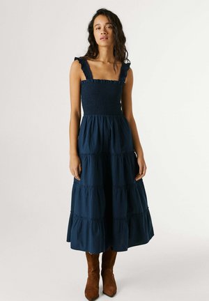 Žena stojí oblečená v bezrukávových námorníckych modrých vrstvených šatách s volánovými ramienkami a hnedými semišovými čižmami na svetlom jednofarebnom pozadí.