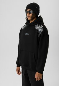 Zwarte hoodie met witte bloemenborduring op de schouders, vooraan een kangoeroezak en ribgebreide manchetten. Draagt een zwarte beanie en zonnebril.