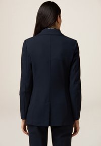 Femme aux longs cheveux foncés portant un blazer bleu marine cintré et un pantalon assorti, debout de dos sur un fond beige uni.