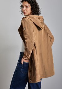 Lekka, beżowa kurtka z kapturem, z elastycznymi mankietami i naszywką z logo na ramieniu, noszona na kremowej bluzce i niebieskich dżinsach.