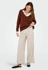 Pull en tricot marron avec un col en V, associé à un pantalon en velours côtelé beige clair à jambes larges avec des plis devant et un détail de bouton noir. Chaussures noires.