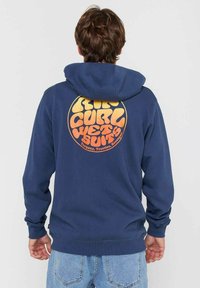 Persoon in een marineblauwe hoodie met een oranje cirkelvormig "Rip Curl Wetsuits" logo op de rug, in combinatie met lichtblauwe jeans, staande met de rug naar ons toegekeerd.