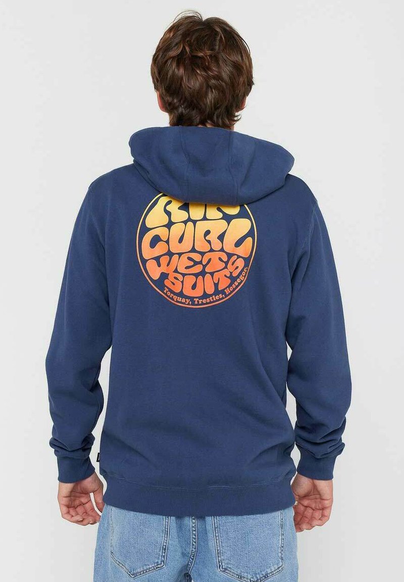 Persona vestida con una sudadera azul marino con un logo circular naranja de "Rip Curl Wetsuits" en la espalda, combinada con jeans de color azul claro, de pie y de espaldas.